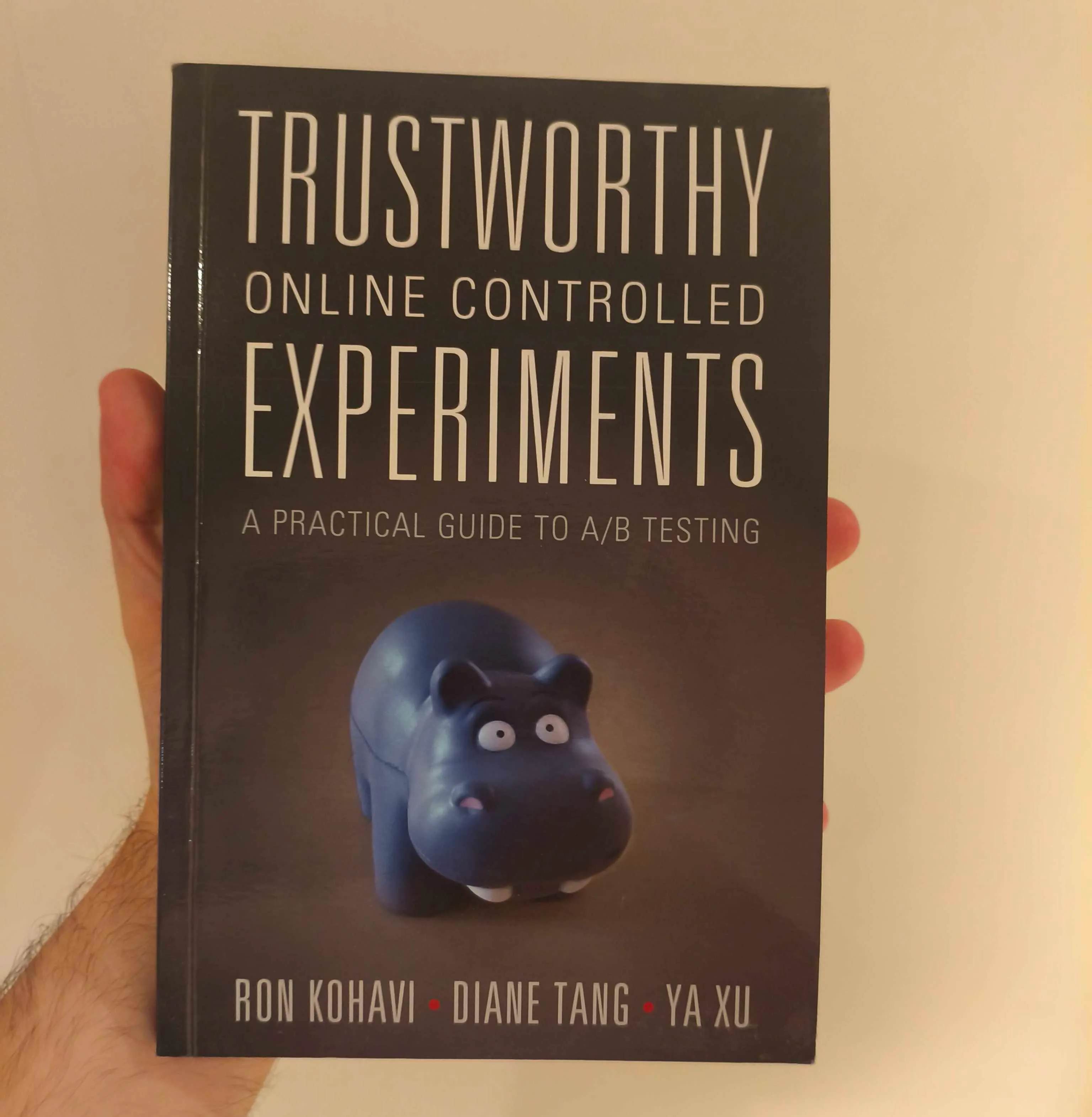 A hand holding a paperback copy of Trustworthy Online Controlled Experiments by Kohavi, Tang and Xu, the cover showing a small blue toy hippo